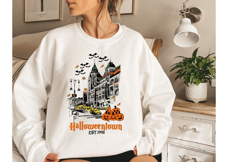 Puede incluir: Sudadera blanca con un estampado gr&aacute;fico de un horizonte urbano con el texto "Halloweentown Est. 1998" y una pila de calabazas.