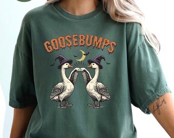 May include: A moss green t-shirt with the word "GOOSEBUMPS" in orange lettering. The shirt features a graphic of two geese wearing witch hats, with a crescent moon and bats. The shirt is a comfortable, casual style.
