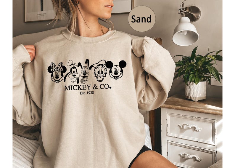 May include: Sand-coloured sweatshirt with a black graphic of Mickey Mouse, Minnie Mouse, Goofy, Pluto, Donald Duck, and Mickey Mouse again. The text "MICKEY & CO." and "Est. 1928" are printed below the characters. The sweatshirt is long-sleeved.