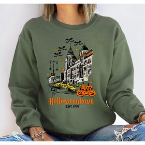 Puede incluir: Una sudadera verde con un estampado gr&aacute;fico de un horizonte de ciudad con un edificio que dice "Halloweentown Est. 1998" y una pila de calabazas.