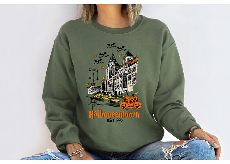 Puede incluir: Una sudadera verde con un estampado gr&aacute;fico de un horizonte de ciudad con un edificio que dice "Halloweentown Est. 1998" y una pila de calabazas.