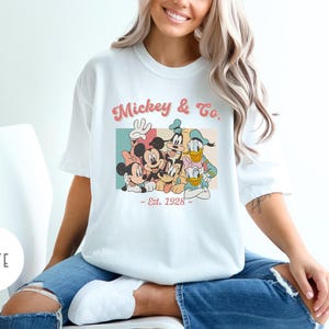 May include: White t-shirt with a vintage-style graphic of Mickey Mouse and friends. The graphic features Mickey Mouse, Minnie Mouse, Donald Duck, Goofy, and Pluto. The text on the shirt reads "Mickey & Co. - Est. 1928 - "