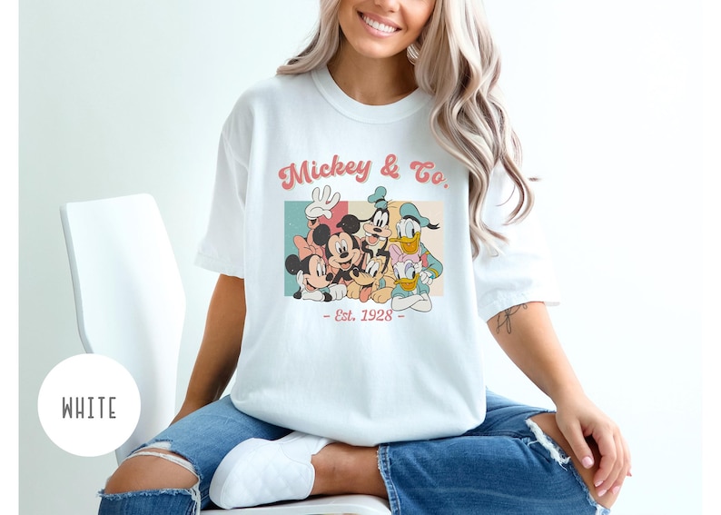 May include: White t-shirt with a vintage-style graphic of Mickey Mouse and friends. The graphic features Mickey Mouse, Minnie Mouse, Donald Duck, Goofy, and Pluto. The text on the shirt reads "Mickey & Co. - Est. 1928 - "
