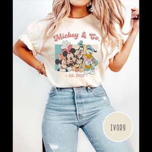 May include: A white t-shirt with a vintage-style graphic of Mickey Mouse, Minnie Mouse, Donald Duck, and Goofy. The graphic says "Mickey & Co. - Est. 1928". The shirt is being modeled by a person wearing blue jeans. The color of the shirt is ivory.