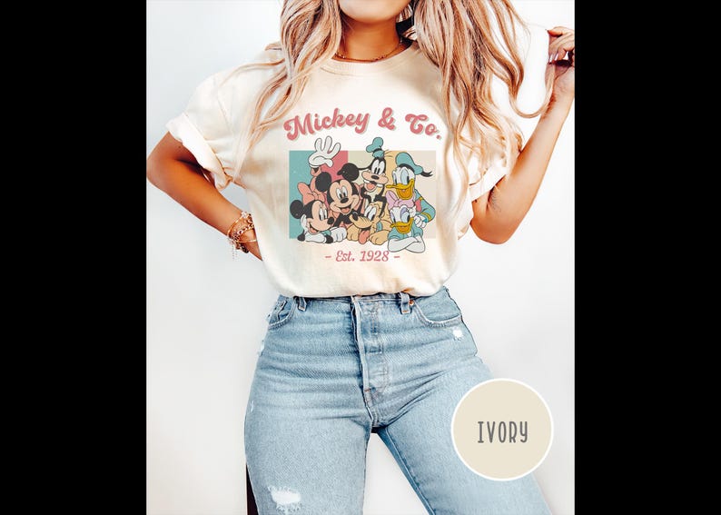 May include: A white t-shirt with a vintage-style graphic of Mickey Mouse, Minnie Mouse, Donald Duck, and Goofy. The graphic says "Mickey & Co. - Est. 1928". The shirt is being modeled by a person wearing blue jeans. The color of the shirt is ivory.