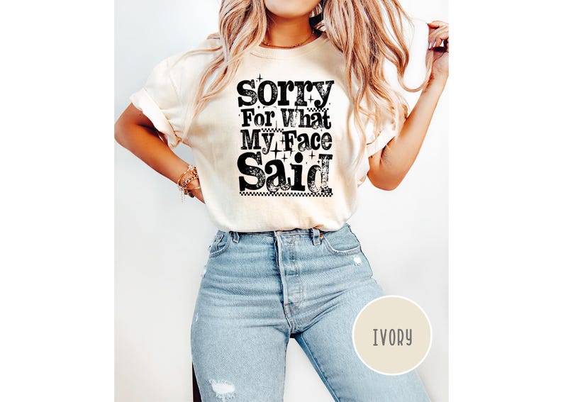 May include: Ivory-colored t-shirt with the text "Sorry For What My Face Said" in a distressed black font. The shirt is paired with light blue jeans. The overall style is casual and relaxed.