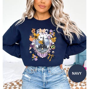 May include: Navy blue sweatshirt featuring a Halloween-themed design. The graphic includes a castle, pumpkins, and character-themed elements within an oval frame. The sweatshirt has a crew neck and long sleeves.