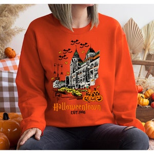 Puede incluir: Sudadera naranja con una ilustraci&oacute;n en blanco y negro de un horizonte de ciudad con murci&eacute;lagos volando por encima. El texto "Halloweentown Est. 1998" est&aacute; impreso debajo de la ilustraci&oacute;n.