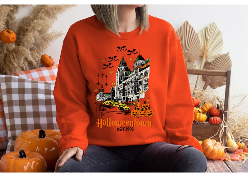 Puede incluir: Sudadera naranja con una ilustraci&oacute;n en blanco y negro de un horizonte de ciudad con murci&eacute;lagos volando por encima. El texto "Halloweentown Est. 1998" est&aacute; impreso debajo de la ilustraci&oacute;n.