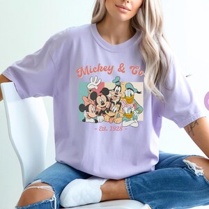May include: A lavender t-shirt with a vintage-style graphic of Mickey Mouse and friends. The graphic features Mickey, Minnie, Donald, Goofy, and Pluto. The text on the shirt reads "Mickey & Co. - Est. 1928 - "