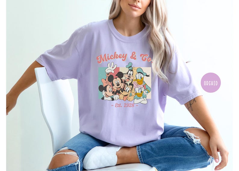 May include: A lavender t-shirt with a vintage-style graphic of Mickey Mouse and friends. The graphic features Mickey, Minnie, Donald, Goofy, and Pluto. The text on the shirt reads "Mickey & Co. - Est. 1928 - "