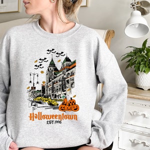 Puede incluir: Sudadera gris con un estampado gr&aacute;fico de un horizonte de ciudad con murci&eacute;lagos, calabazas y el texto "Halloweentown Est. 1998".