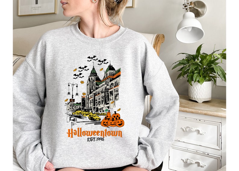 Puede incluir: Sudadera gris con un estampado gr&aacute;fico de un horizonte de ciudad con murci&eacute;lagos, calabazas y el texto "Halloweentown Est. 1998".
