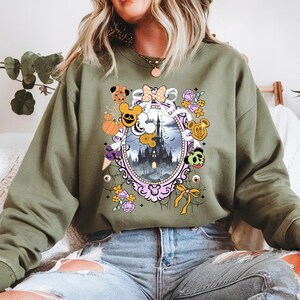 May include: Olive green sweatshirt featuring a Halloween-themed graphic. The design includes a haunted castle within a decorative frame, surrounded by cartoon characters, pumpkins, and floral elements. The sweatshirt is made of a soft material.