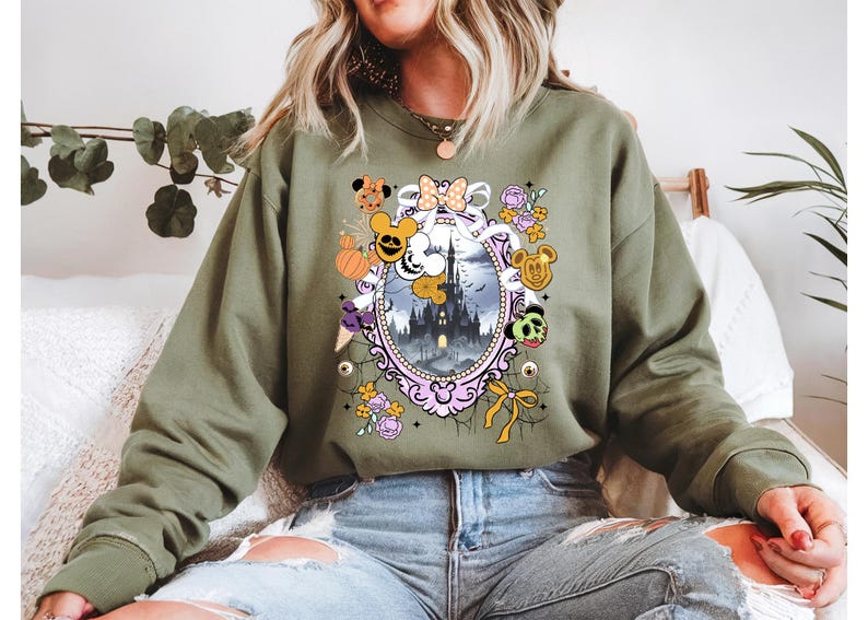 May include: Olive green sweatshirt featuring a Halloween-themed graphic. The design includes a haunted castle within a decorative frame, surrounded by cartoon characters, pumpkins, and floral elements. The sweatshirt is made of a soft material.
