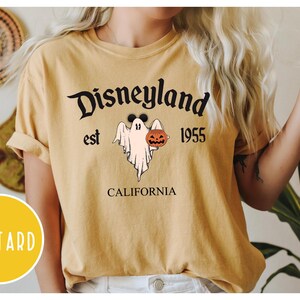 Op de afbeelding: Een mosterdgeel T-shirt met een afbeelding van een spook dat een jack-o-lantern vasthoudt. De tekst "Disneyland est 1955 Californi&euml;" staat op het T-shirt gedrukt.