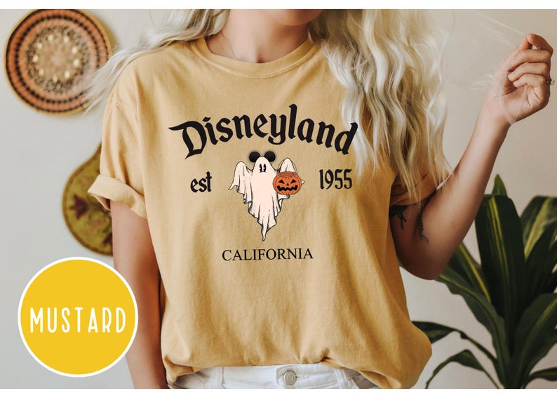 Op de afbeelding: Een mosterdgeel T-shirt met een afbeelding van een spook dat een jack-o-lantern vasthoudt. De tekst "Disneyland est 1955 Californi&euml;" staat op het T-shirt gedrukt.