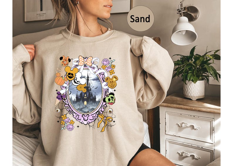 May include: Sand-colored sweatshirt featuring a Halloween-themed graphic. The design includes a castle, pumpkins, Mickey Mouse-inspired characters, and floral elements within an ornate oval frame. The overall aesthetic is whimsical and festive.