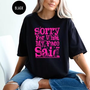 May include: Black t-shirt with the text "Sorry For What My Face Said" in bright pink. The text has a distressed, vintage look. The shirt is a casual, oversized fit.