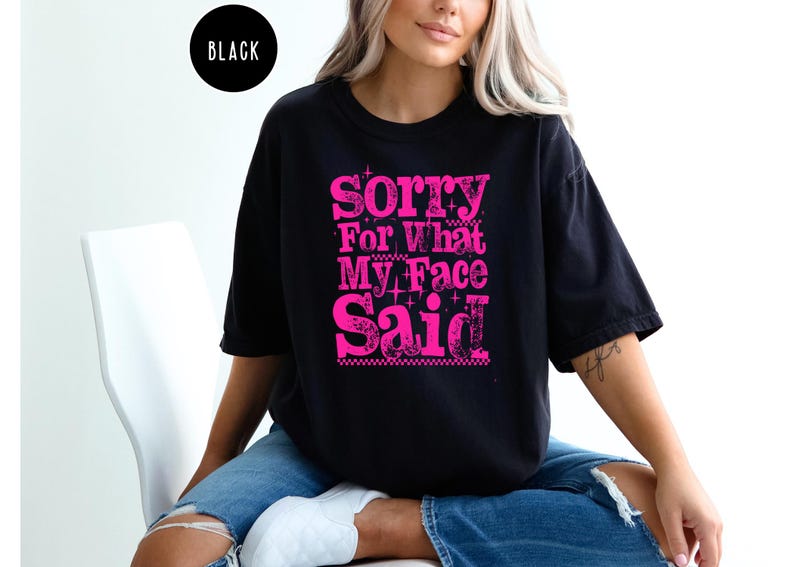 May include: Black t-shirt with the text "Sorry For What My Face Said" in bright pink. The text has a distressed, vintage look. The shirt is a casual, oversized fit.