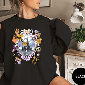 May include: Black sweatshirt featuring a Halloween-themed design. The design includes a castle, Mickey Mouse characters, pumpkins, and floral elements within an oval frame. The sweatshirt has long sleeves and a round neckline.