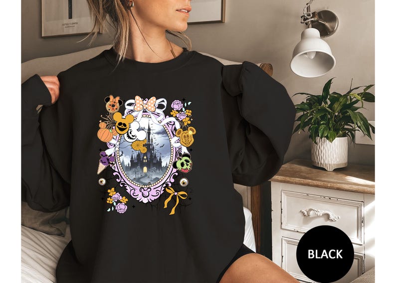 May include: Black sweatshirt featuring a Halloween-themed design. The design includes a castle, Mickey Mouse characters, pumpkins, and floral elements within an oval frame. The sweatshirt has long sleeves and a round neckline.