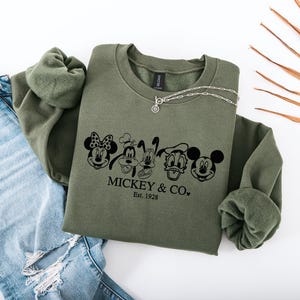 May include: An olive green sweatshirt with a crew neck and long sleeves. The front features a black graphic of Mickey Mouse, Minnie Mouse, Goofy, Donald Duck, and Mickey Mouse again, with the text "MICKEY & CO. Est. 1928". A silver necklace rests on the sweatshirt.