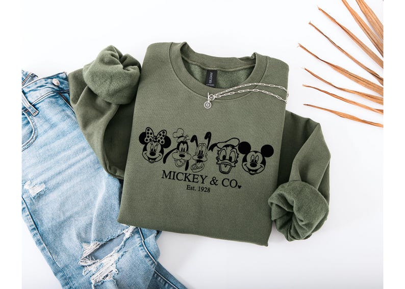 May include: An olive green sweatshirt with a crew neck and long sleeves. The front features a black graphic of Mickey Mouse, Minnie Mouse, Goofy, Donald Duck, and Mickey Mouse again, with the text "MICKEY & CO. Est. 1928". A silver necklace rests on the sweatshirt.