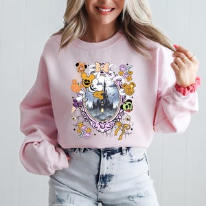 May include: A light pink sweatshirt featuring a framed illustration of a castle with cartoon characters and Halloween-themed elements. The design includes pumpkins, bows, and character faces. The sweatshirt has long sleeves.