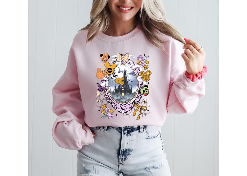 May include: A light pink sweatshirt featuring a framed illustration of a castle with cartoon characters and Halloween-themed elements. The design includes pumpkins, bows, and character faces. The sweatshirt has long sleeves.