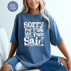 May include: A blue jean-colored t-shirt with the text "Sorry For What My Face Said" in white, decorative lettering. The shirt is a short-sleeved, casual style. A circular "Blue Jean" label is in the upper left corner.