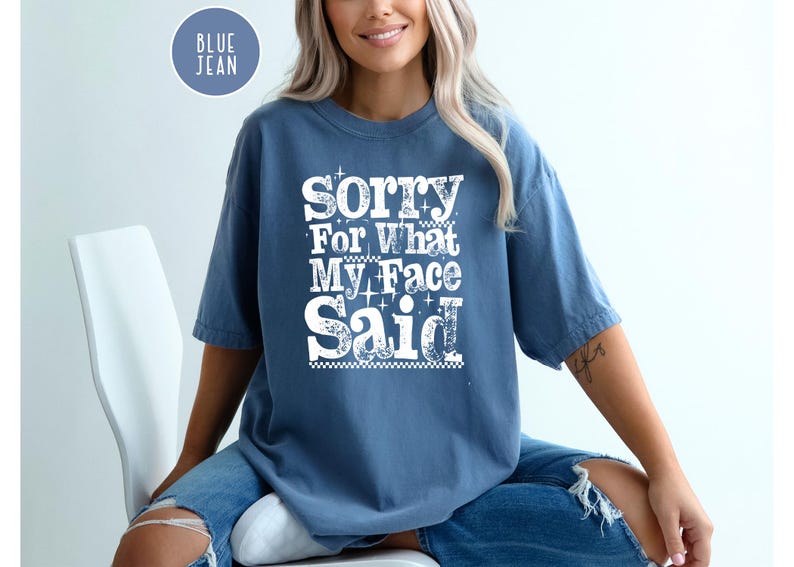 May include: A blue jean-colored t-shirt with the text "Sorry For What My Face Said" in white, decorative lettering. The shirt is a short-sleeved, casual style. A circular "Blue Jean" label is in the upper left corner.