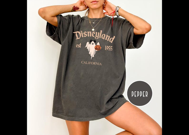 Op de afbeelding: Een donkergrijs T-shirt met een grafische print van een spook dat een jack-o-lantern vasthoudt. De tekst "Disneyland est 1955 Californi&euml;" staat op het T-shirt gedrukt.