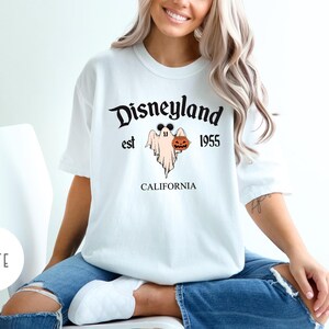 Op de afbeelding: Wit T-shirt met een zwarte afbeelding van een geest die een pompoen vasthoudt. De afbeelding zegt "Disneyland est 1955 Californi&euml;".