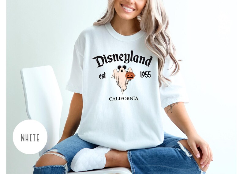 Op de afbeelding: Wit T-shirt met een zwarte afbeelding van een geest die een pompoen vasthoudt. De afbeelding zegt "Disneyland est 1955 Californi&euml;".