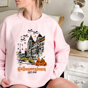 Puede incluir: Sudadera rosa con un estampado gr&aacute;fico de un horizonte de ciudad con murci&eacute;lagos, calabazas y el texto "Halloweentown Est. 1998".