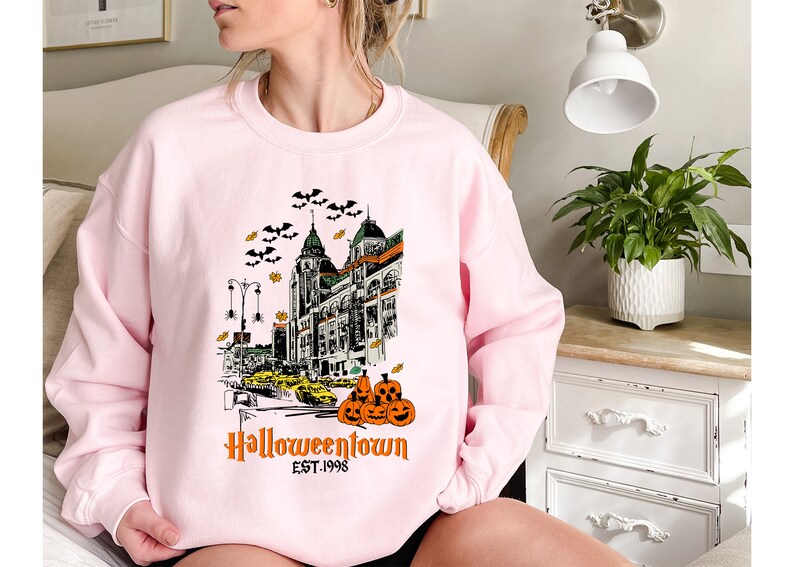 Puede incluir: Sudadera rosa con un estampado gr&aacute;fico de un horizonte de ciudad con murci&eacute;lagos, calabazas y el texto "Halloweentown Est. 1998".