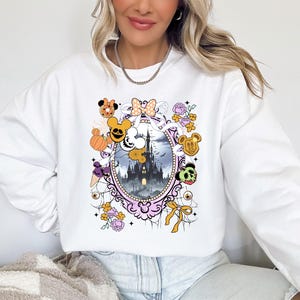 May include: White sweatshirt featuring a colorful graphic of a spooky castle scene within a decorative frame. The design includes cartoon characters, pumpkins, and floral accents, creating a festive Halloween theme.
