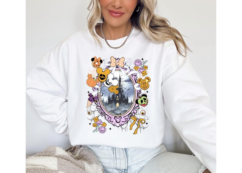 May include: White sweatshirt featuring a colorful graphic of a spooky castle scene within a decorative frame. The design includes cartoon characters, pumpkins, and floral accents, creating a festive Halloween theme.