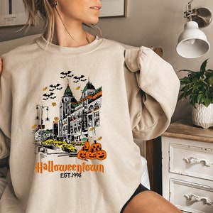 Puede incluir: Una sudadera beige con un estampado gr&aacute;fico de un horizonte de ciudad con calabazas y murci&eacute;lagos. El texto "Halloweentown Est. 1998" est&aacute; impreso debajo del gr&aacute;fico.