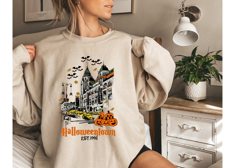 Puede incluir: Una sudadera beige con un estampado gr&aacute;fico de un horizonte de ciudad con calabazas y murci&eacute;lagos. El texto "Halloweentown Est. 1998" est&aacute; impreso debajo del gr&aacute;fico.