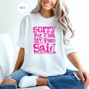 May include: White t-shirt with the text "Sorry For What My Face Said" in bright pink. The shirt is a crew neck style and appears to be made of a soft material. The text is in a bold, stylized font.