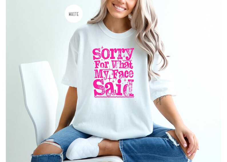May include: White t-shirt with the text "Sorry For What My Face Said" in bright pink. The shirt is a crew neck style and appears to be made of a soft material. The text is in a bold, stylized font.