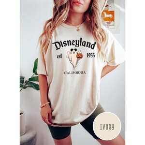Op de afbeelding: Een cr&egrave;mekleurig T-shirt met een zwarte afbeelding van een geest die een pompoen vasthoudt. De tekst "Disneyland est 1955 CALIFORNI&Euml;" staat op het T-shirt gedrukt.