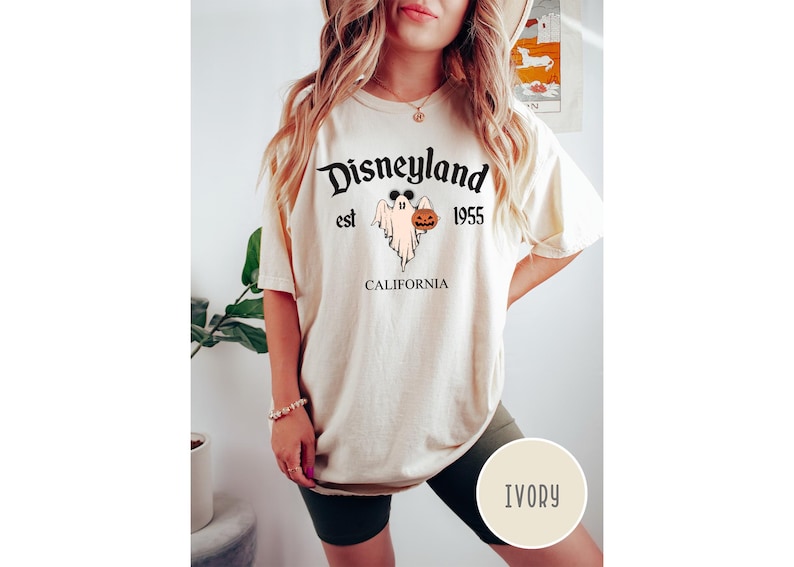 Op de afbeelding: Een cr&egrave;mekleurig T-shirt met een zwarte afbeelding van een geest die een pompoen vasthoudt. De tekst "Disneyland est 1955 CALIFORNI&Euml;" staat op het T-shirt gedrukt.