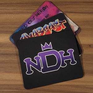 May include: A stack of square mousepads with various designs. The top mousepad is black with a purple and white "NDH" logo and a crown. The second mousepad has the word "ANABAST" in a graffiti style. The third mousepad has the text "NED OUT MERCH".