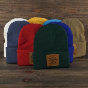 May include: A collection of colorful beanies in white, navy, red, green, blue, yellow, and tan. Each beanie has a folded brim and a brown leather patch with the text "Broadway" and a logo.