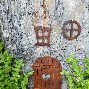 May include: A rusty metal fairy door with a heart-shaped window, a rusty metal window, and a rusty metal circle with a design inside. The door is attached to a tree trunk.