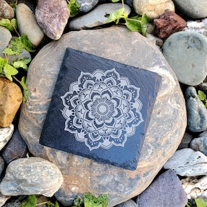May include: A black slate coaster with a silver mandala design. The coaster is sitting on a gray rock.