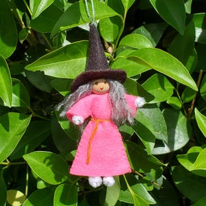 May include: A hanging ornament featuring a doll with a pointed dark brown felt hat, gray yarn hair, and a bright pink felt dress. The doll has a simple painted face, a thin orange waist tie, and small white pom-pom accents at the hands and feet.